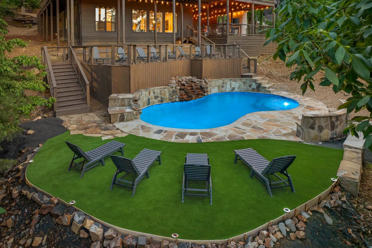 A client cabin we service at dusk — multi-level decks with string lights above a stone-edged pool and waterfall