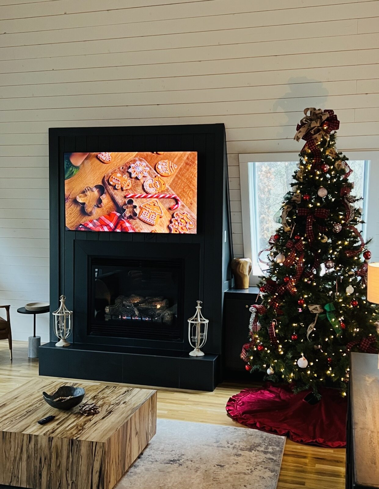 A decorated Christmas tree with plaid ribbon beside a black fireplace surround in a warm, wood-panelled cabin living room