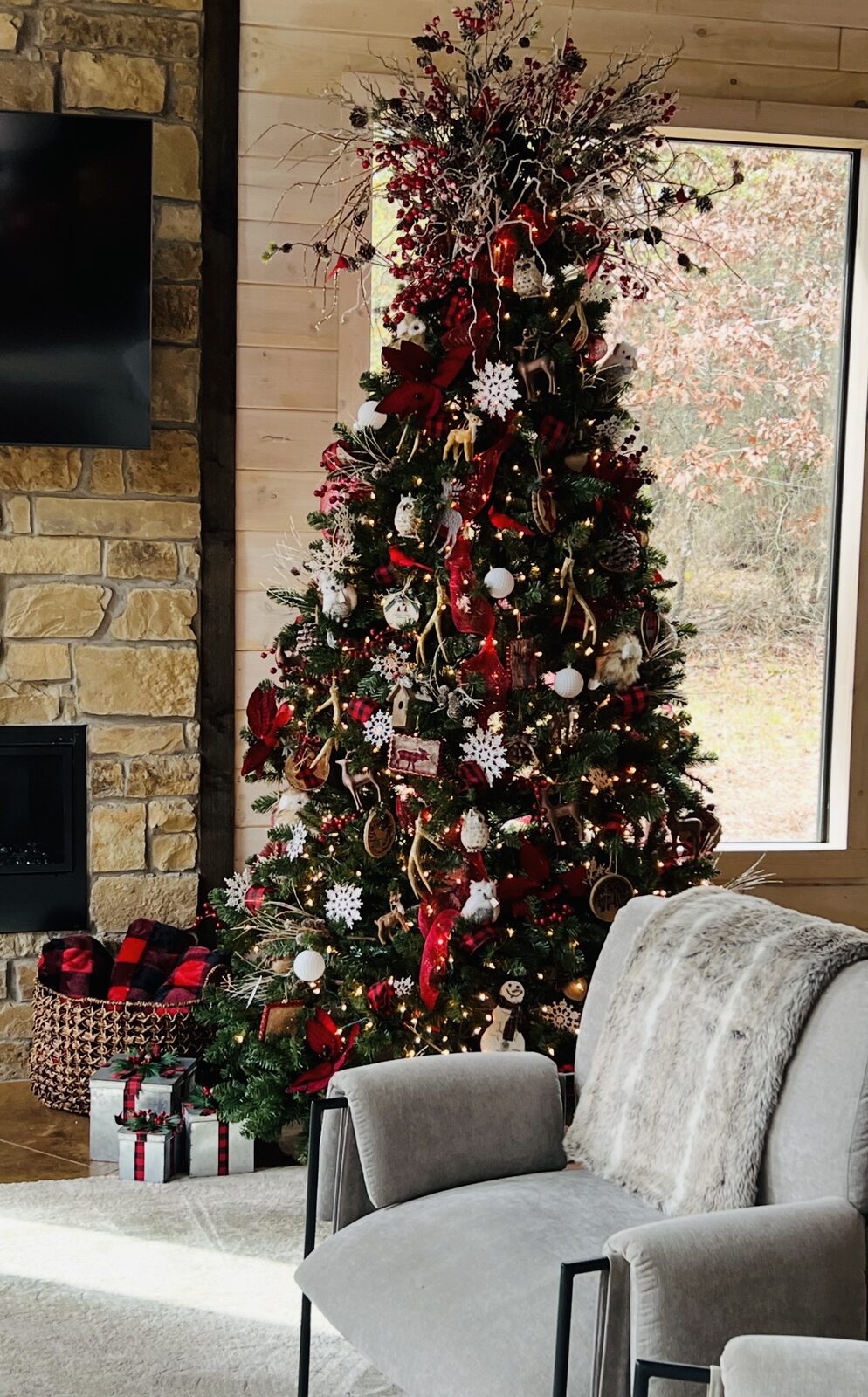 A fully decorated Christmas tree beside a stone fireplace in a cabin, trimmed with red and plaid ribbon, wrapped gifts underneath