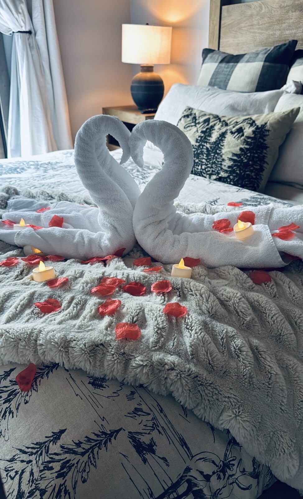 Two white towels folded into swans forming a heart on a bed with red rose petals and flameless tea lights