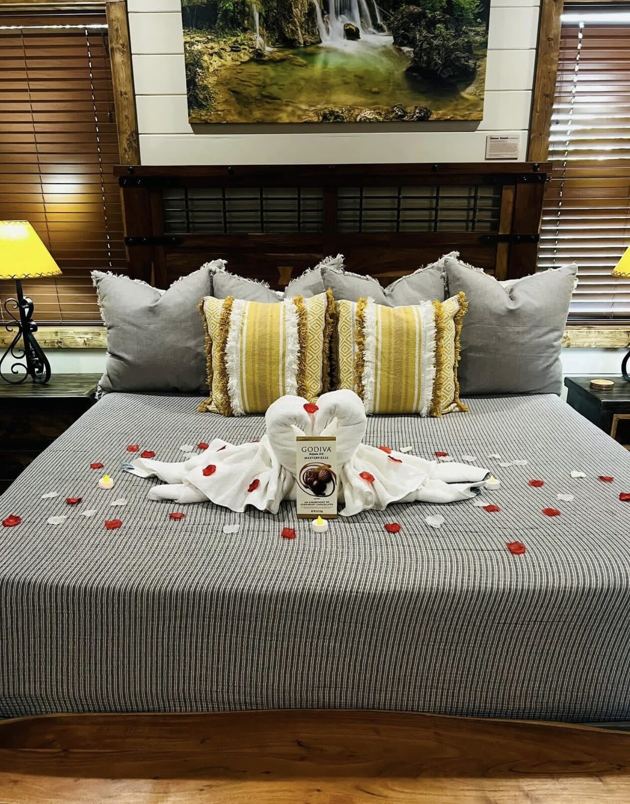 King bed styled with towel swans, a Godiva chocolate box and rose petals beneath a large framed waterfall photograph